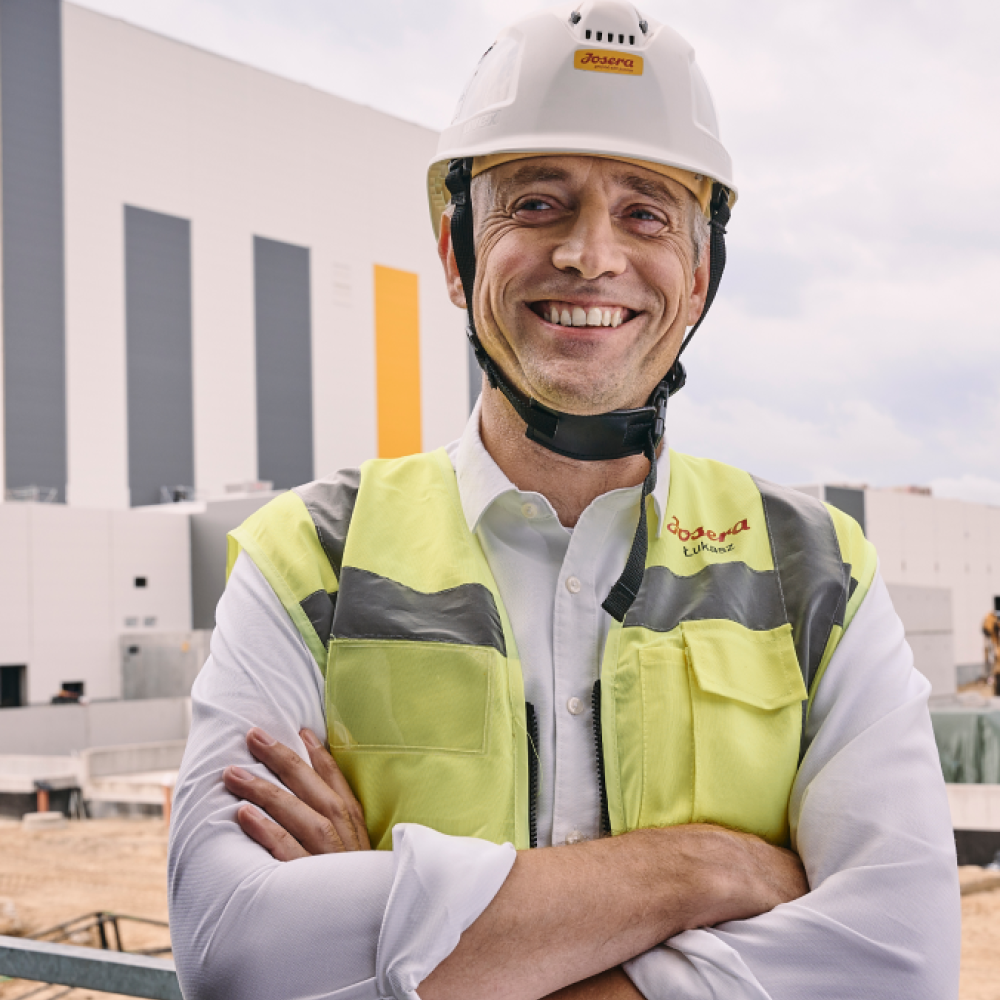 Ein Mann verschränkt die Arme und lacht in die Kamera. Er hat einen weißen Helm mit Joser aLogo auf und eine gelbe Warnweste an. Er steht draußen auf der Baustelle, im  Hintergrund das neue Werk.