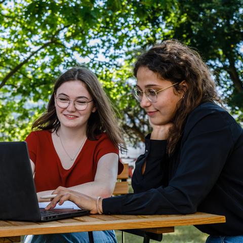Zwei Mitarbeiterinnen sitzen gemeinsam im Park und schauen auf den Laptop.